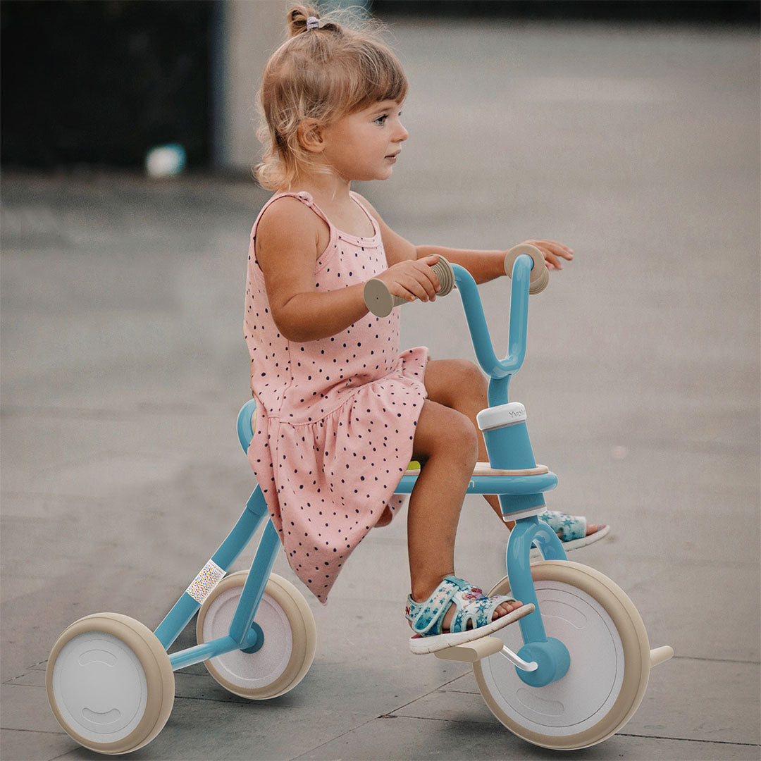 Een peuter meisje in een roze jurk rijdt buiten op de blauwe driewieler.