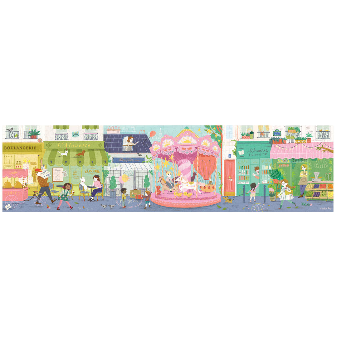 Volledige Moulin Roty panoramapuzzel met een lange, kleurrijke straatscène met winkels, een draaimolen en figuren.