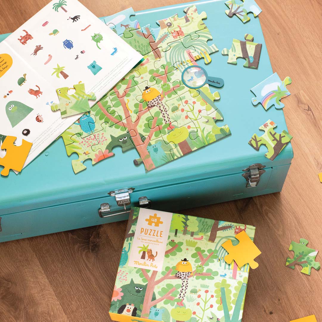 Verspreide stukjes van de Moulin Roty legpuzzel 'Prachtige aarde' liggen naast de doos en een zoekboekje op een houten ondergrond.