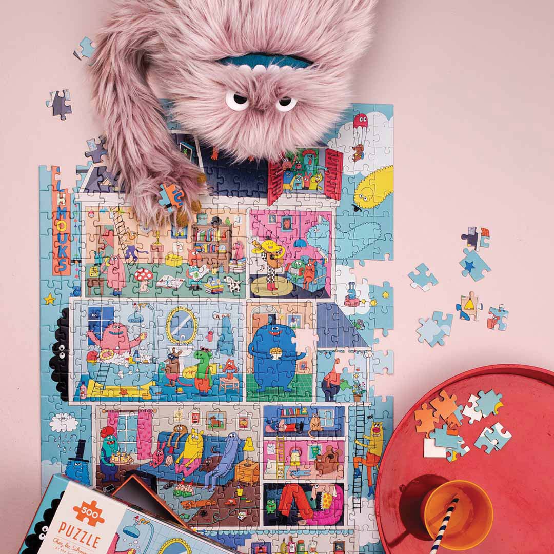 Een deels gelegde Moulin Roty puzzel van een monsterhuis, met een roze pluizig monster speelgoed erop en losse puzzelstukjes.