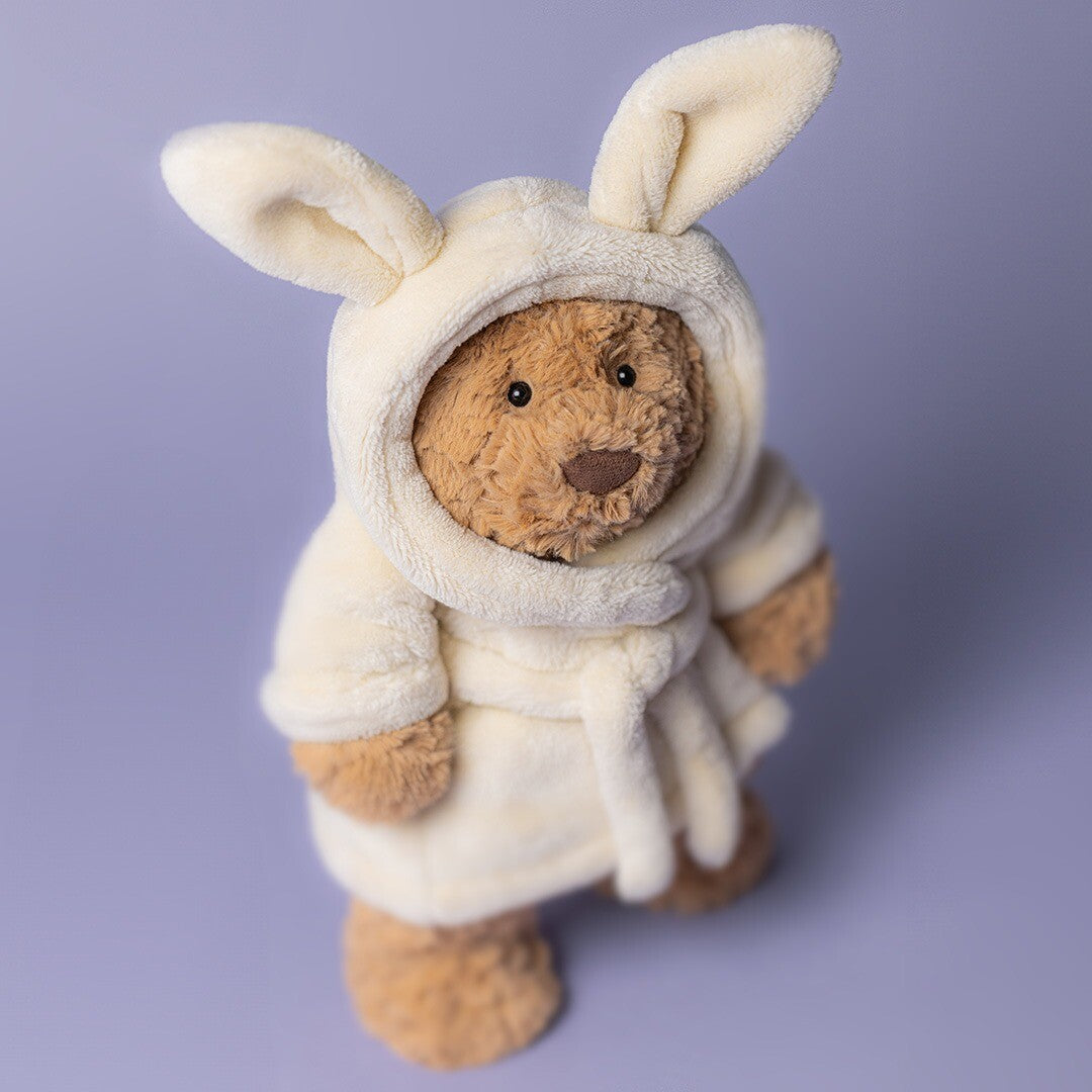 Jellycat knuffelbeer Bartholomew in een crèmekleurige badjas met konijnenoren, staand op een paarse achtergrond.