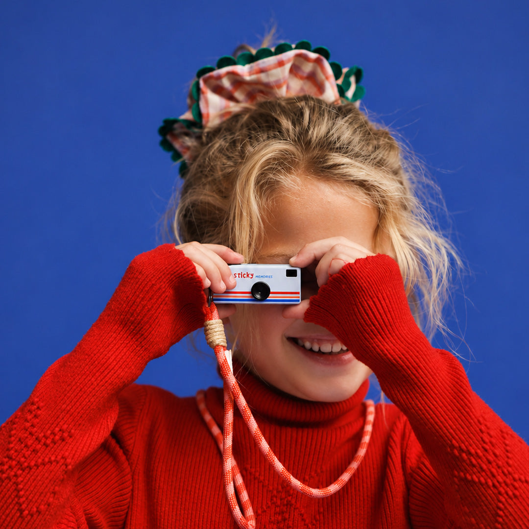 Een kind met een rode trui poseert met de blauwe Sticky Lemon mini kindercamera voor het oog.